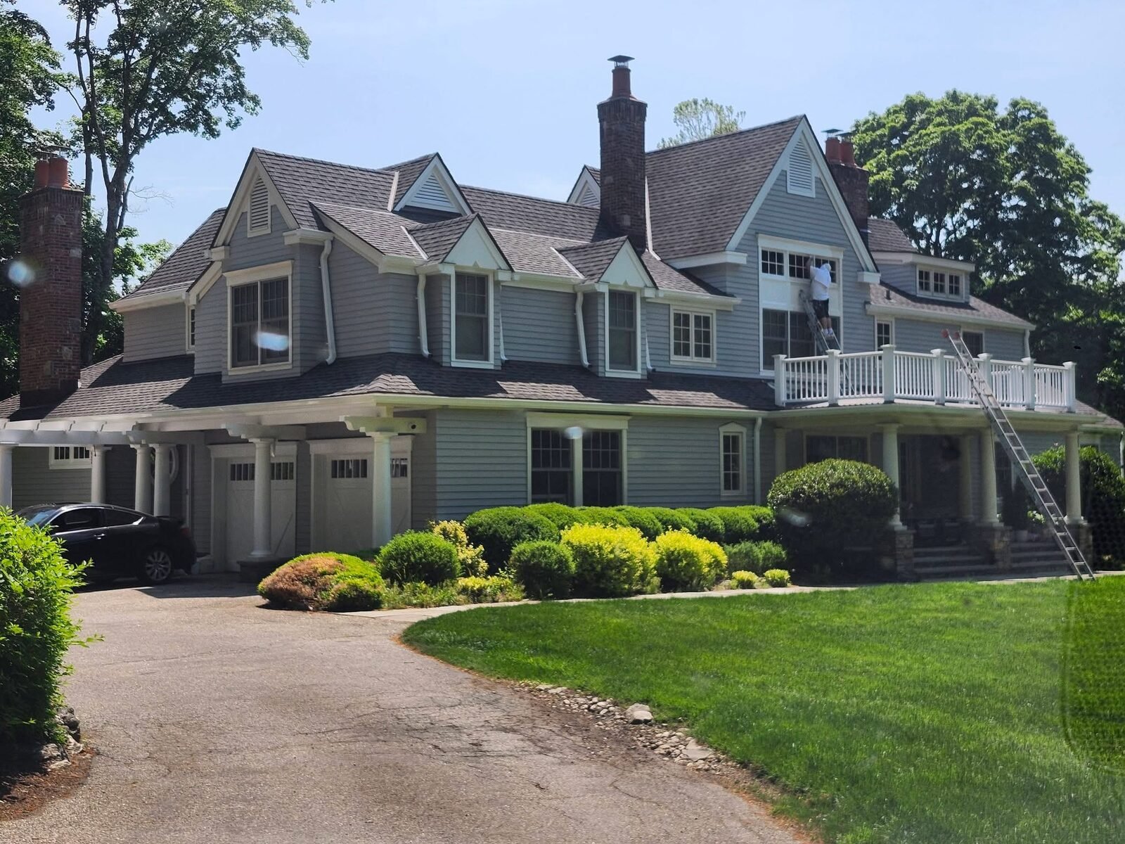 Painter on a ladder repainting upper window trim on a large gray shingle home